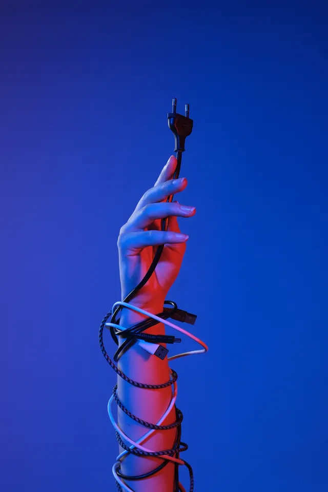 Hand holding an electrical plug, wrapped in cables and lit blue and red, photographed by Edith van Aken
